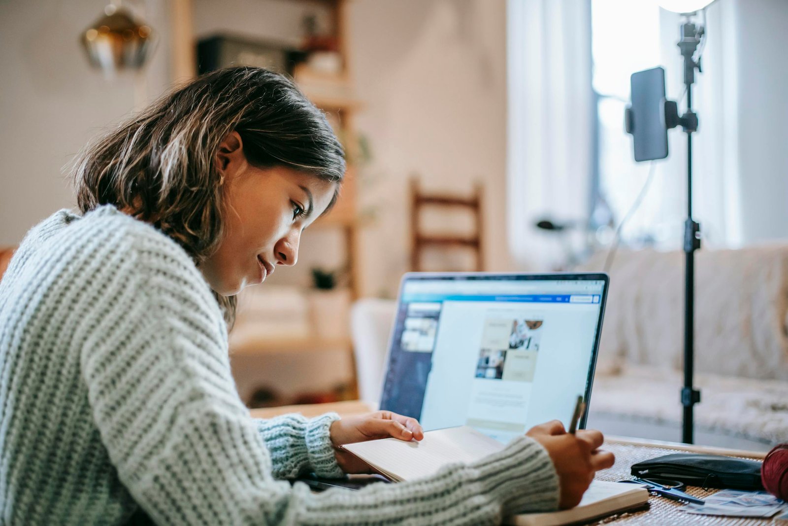 Side view of concentrated young Latin American female freelancer in casual clothes using laptop while taking notes in notebook with pen at table near ring light with phone in room